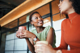 Two stylish women in a modern office setting share a lively conversation over coffee. Soft natural light highlights their expressions and attire.